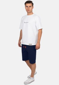 Witte katoenen t-shirt met zwart logo op de borst, gecombineerd met marineblauwe shorts met oranje trekkoorden. Model draagt witte sneakers.