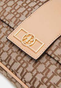 Detailní záběr na béžovou a hnědou vzorovanou kabelku se zlatým obdélníkovým a kulatým logem na uzávěru z hladké kůže.