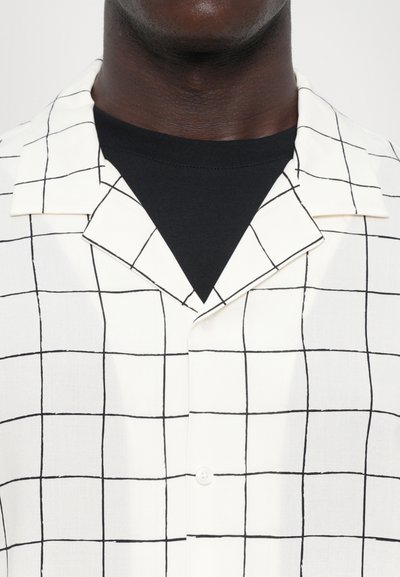 Witte korte mouwen shirt met een zwart ruitpatroon, knoopsluiting en een geknipt kraag, gelaagd over een zwart t-shirt met ronde hals.