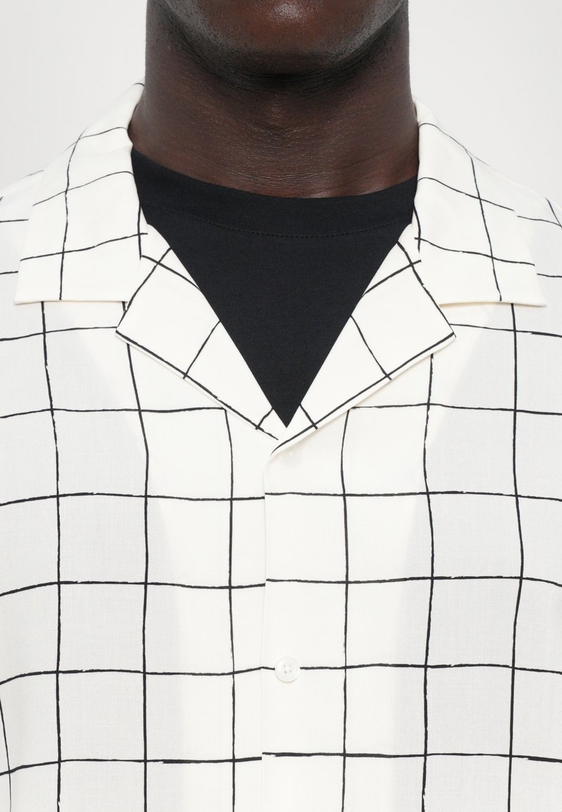 Vit kortärmad skjorta med ett svart rutmönster, knäppning fram och en skårad krage, lagd över en svart t-shirt med rund halsringning.