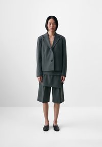 Gray tailored blazer with a notched collar, single button closure; layered gray shorts with a structured silhouette; black slip-on shoes.