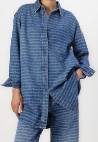 Farkkupaita, jossa sininen tausta ja toistuva vaaleansininen tekstikuosi, napitettava etuosa ja ylivuotava malli. Pitkät hihat näkyvissä.