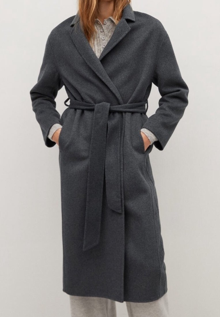 Femme portant un manteau en laine gris foncé ceinturé par-dessus un pull boutonné gris clair et un pantalon gris clair, les mains dans les poches du manteau.