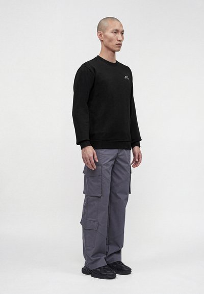 Άνδρας που στέκεται φορώντας μαύρο μακρυμάνικο φούτερ, γκρι παντελόνι cargo και μαύρα αθλητικά παπούτσια σε απλό ανοιχτό φόντο.