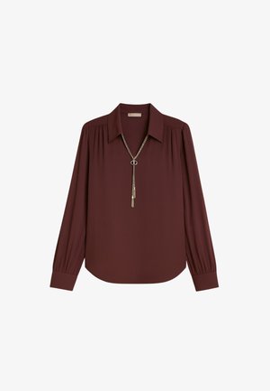 Blusa bordeaux a maniche lunghe con colletto a punta e polsini plissettati, caratterizzata da una collana decorativa in argento con un pendente circolare.