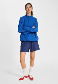 Blå atletisk jacka med vit kantning, tillsammans med marina shorts, knähöga vita strumpor och röda sneakers. Har en avslappnad passform och dragkedjeförslutning.