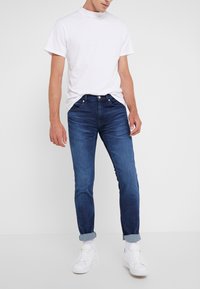 Jeans de mezclilla azul oscuro con un corte ajustado, que presentan un diseño clásico de cinco bolsillos y dobladillos doblados, combinados con una camiseta blanca lisa y zapatillas blancas.