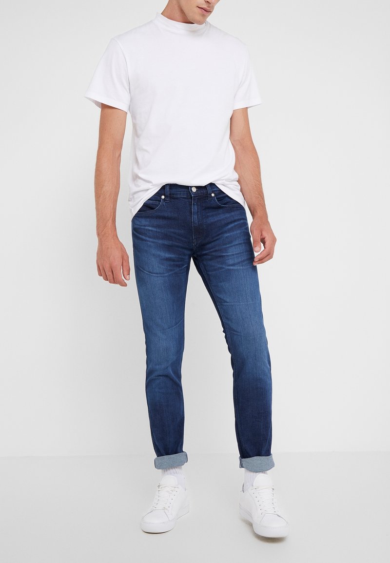 Jeans de mezclilla azul oscuro con un corte ajustado, que presentan un diseño clásico de cinco bolsillos y dobladillos doblados, combinados con una camiseta blanca lisa y zapatillas blancas.