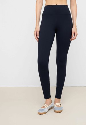 Kvinde iført sorte leggings med høj talje og lyseblå balletflade sko med krydsremme, stående mod en ensfarvet hvid baggrund.