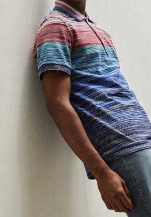 Mann trägt ein kurzärmeliges Hemd mit Kragen und horizontalen Streifen in Rot, Grün, Blau und Marineblau, lehnt an einer hellen Wand.