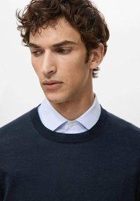 Jonge man met krullend bruin haar, gekleed in een marineblauwe trui over een lichtblauw overhemd met kraag, kijkt naar beneden en opzij tegen een effen achtergrond.