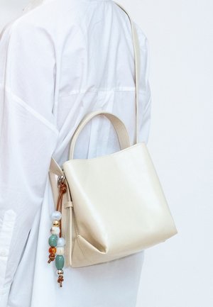 Bolso de cuero color crema con asas superiores y correa para el hombro, llevado sobre camisa blanca de manga larga, adornado con un charm de borla de cuentas de colores.