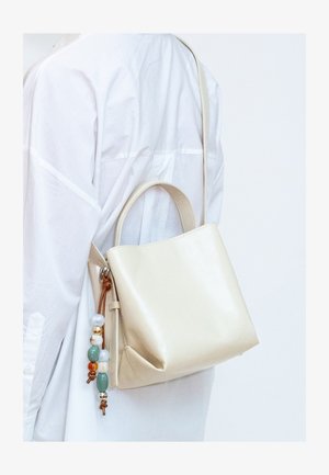 Sac à main en cuir crème avec poignées supérieures et bandoulière, porté sur une chemise blanche à manches longues, orné d'un pompon décoratif coloré en perles.