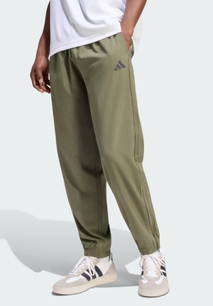Person trägt olivgrüne Adidas-Trainingshose und weiße Sneakers mit schwarzen Streifen, steht mit einer Hand in der Tasche.