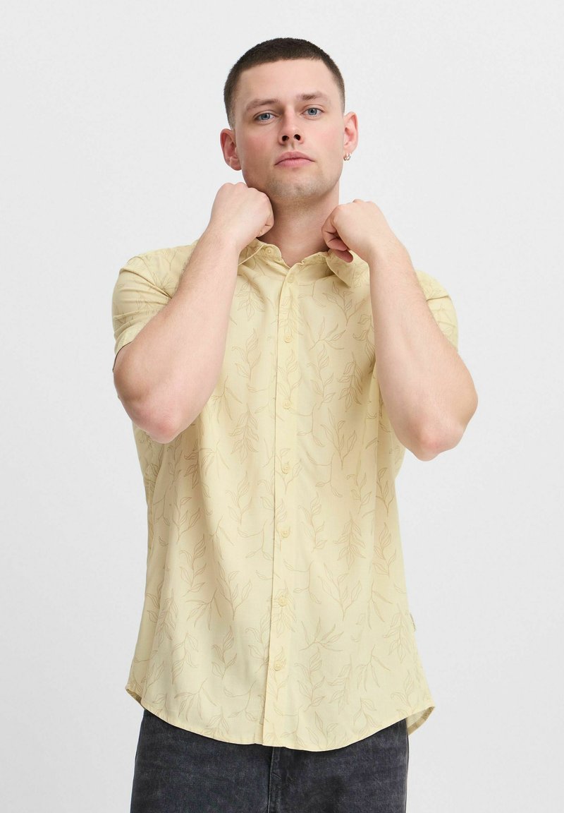 Chemise à manches courtes en tissu jaune clair avec un motif de feuilles subtil. Présente une fermeture à boutons sur le devant et un ourlet légèrement incurvé.