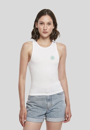 Jeune femme portant un haut blanc sans manches avec un petit motif de fleur verte souriante et un short en jean bleu clair.