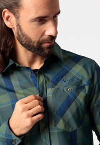 Camisa de cuadros verde y azul con botones y un bolsillo en el pecho, hecha de una tela suave, con un logo bordado en la parte superior izquierda.