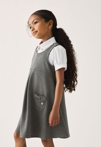 Gekleidet mit einem grauen Baumwollkleid im ausgestellten Design, das eine Fronttasche mit einer Schleife und einem Herzanhänger hat. Über einem weißen Hemd mit Kragen getragen.