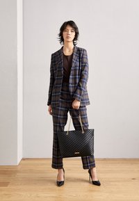 Plaid suit in navy and brown with a fitted jacket and wide-leg trousers. Black quilted handbag with chain handle. High black heels.