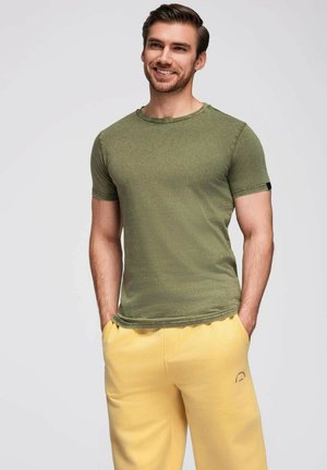 Olivgrön kortärmad t-shirt med rund halsringning, kombinerad med gula sweatpants. T-shirten har en något texturerad yta och en avslappnad passform.