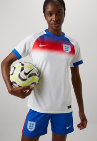 Equipación de fútbol de Nike con una camiseta blanca que presenta un degradado rojo y acentos azules, acompañada de pantalones cortos azules que exhiben el escudo de Inglaterra. Sosteniendo un balón con un diseño.