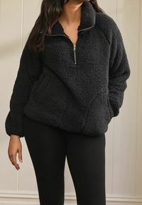 Polaire noire avec col demi-fermeture éclair, manches raglan et poche avant. Texture douce, coupe décontractée, adaptée à la superposition.