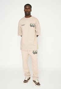 Camiseta de algodón beige y pantalones de chándal a juego con un ajuste holgado, acentos de logo bordado y una silueta relajada con cinturilla ajustable.