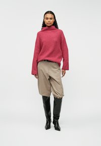 Sieviete, tērpta brīvā sarkanā augstajā kaklā džemperī, bejžās plašās šortos un melnās augstpapēžu zābakos, stāv uz vienkārša fona.