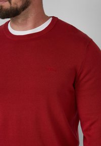 Roter Strickpullover mit Rundhalsausschnitt und langen Ärmeln. Verfügt über ein dezentes, tonales rotes Logo auf der Brust. Glatte Textur.