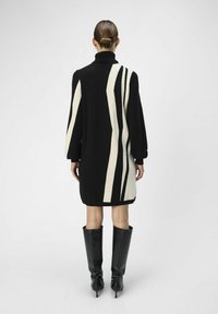 Sort sweaterkjole med høj krave, der har hvide bølgede striber, ribbede manchetter og en løs pasform, parret med knæhøje sorte støvler.