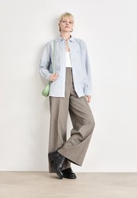 Chemise bleu clair à boutons, haut blanc, pantalon large gris et bottes noires à lacets. Un sac bandoulière vert ajoute une touche de couleur.