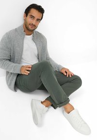 Grijze gebreide cardigan, wit T-shirt, olijfgroene broek en witte sneakers. Model zit in een ontspannen houding tegen een witte achtergrond.