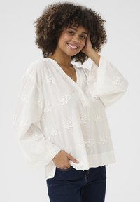 Blusa bianca ricamata con motivi floreali, scollatura a V, maniche lunghe e taglio morbido, abbinata a jeans scuri.