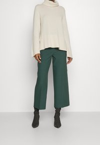 Person i en krämfärgad högkragströja, mörkgröna vida capribyxor och svarta spetsiga stövlar, som står mot en enkel vit bakgrund.