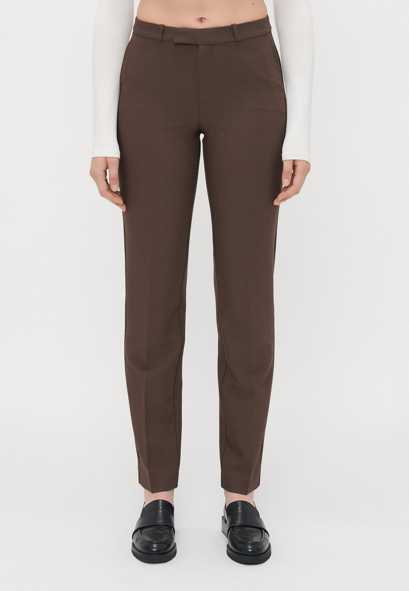 Bruine op maat gemaakte broeken met een gladde textuur, slim fit en een platte voorkant. Gecombineerd met zwarte instapschoenen.