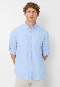 Light blue button-up shirt made of linen with a chest pocket, roll-up sleeves, and a rounded hem. Features simple buttons and a classic collar.