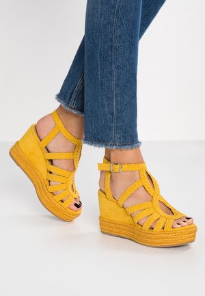 Fødder iført gule flettede kilehælssandaler med ankelstropper kombineret med blå jeans med frynset kant på hvidt gulv.