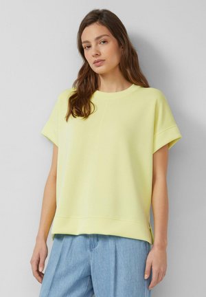 Kobieta o długich brązowych włosach, ubrana w luźną bladożółtą bluzkę z krótkim rękawem i jasnoniebieskie dżinsy, stojąca na jednolitym szarym tle.