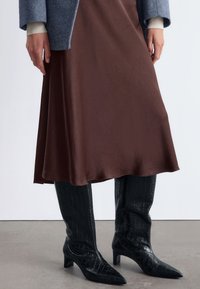 Brauner Satin-Midi-Rock kombiniert mit knielangen schwarzen, krokodilgeprägten Lederstiefeln. Das Outfit hat eine lockere Passform und eine glatte Textur.