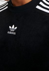 Svart fleece-tröja med rund hals, med tre vita randiga detaljer på axlarna och en vit Adidas-logotyp på bröstet.