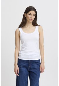 Sieviete ar gariem brūniem matiem, valkājot baltu bezroku topu un zilas džinsas, stāv pret vienkāršu gaišu fonu.