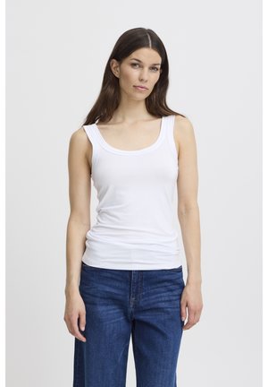 Donna con lunghi capelli castani che indossa una canottiera bianca e jeans blu, in piedi davanti a uno sfondo chiaro e uniforme.