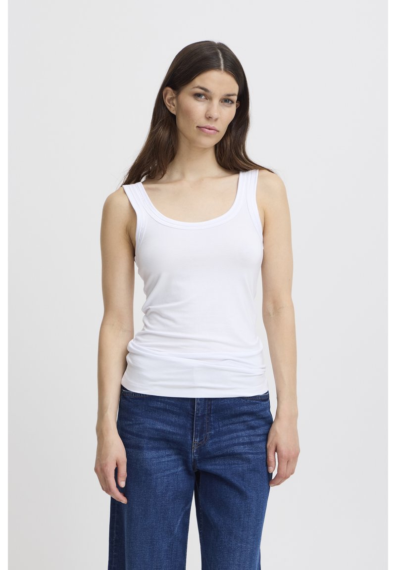 Sieviete ar gariem brūniem matiem, valkājot baltu bezroku topu un zilas džinsas, stāv pret vienkāršu gaišu fonu.