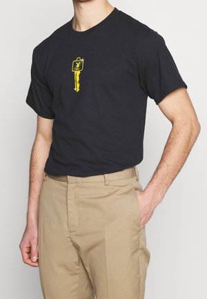 Czarna bawełniana koszulka z nadrukiem żółtego klucza na piersi. Krótkie rękawy i klasyczny krój. W połączeniu z beżowymi spodniami.