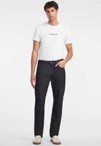 Černé džíny s rovnou siluetou, nošeny s bílým přiléhavým tričkem s logem "GUESS JEANS". Celkový vzhled doplňují casual tenisky.