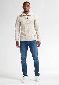 Kermanvärinen palmikkoneulepaita, jossa korkea kaulus ja napitus, yhdistettynä sinisiin farkkuihin ja valkoisiin tennareihin. Tasainen pinta, rento muotoilu.