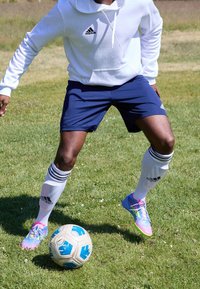 Sweat à capuche blanc avec un logo, short bleu marine, chaussettes blanches à rayures, et crampons multicolores, posés sur un ballon de football texturé sur l'herbe.