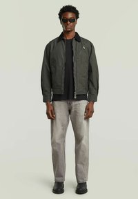 Uomo di colore in giacca a zip verde scuro, maglia nera, jeans grigio chiaro, stivali neri e occhiali da sole neri rettangolari, in piedi su uno sfondo semplice.