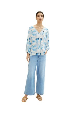 Blouse légère blanche avec des motifs abstraits bleus, présentant un décolleté en V et des manches longues. Associée à un jean large bleu clair et des sandales.