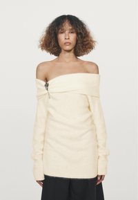 Pull crème décolleté avec une texture douce, des manches longues et un détail de broche en argent décoratif sur le côté gauche.